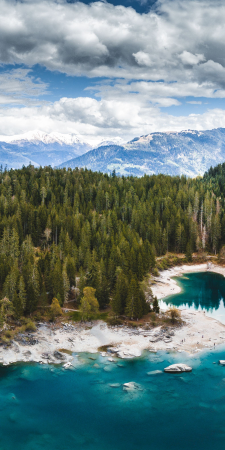 Luftaufnahme eines türkisblauen Bergsees in Graubünden, umgeben von dichten Nadelwäldern und verschneiten Gipfeln unter dramatischen Wolken.