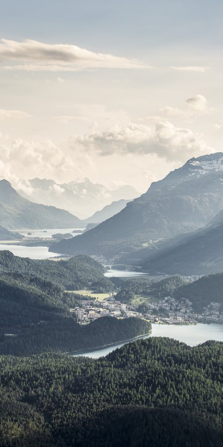 Panoramablick über die Oberengadiner Seen mit glitzernden Gewässern, dichten Wäldern, einer Ortschaft und imposanten Alpengipfeln im Hintergrund.