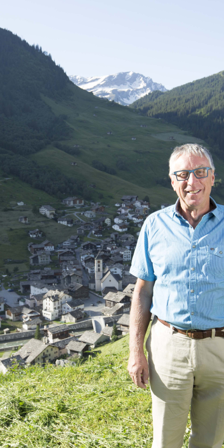 Reto Jörger steht oberhalb des Bergdorfs Vals auf einer sonnigen Wiese, dahinter liegen die traditionellen Häuser und bewaldete Alpenhänge.