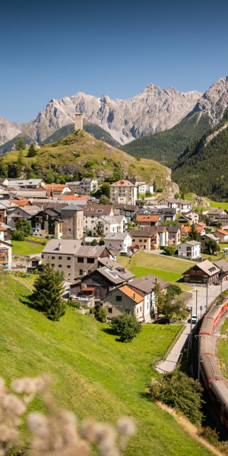Panorama des Engadiner Dorfs Ardez mit der Burgruine Steinsberg, der Rhätischen Bahn, grünen Wiesen und schroffen Berggipfeln.