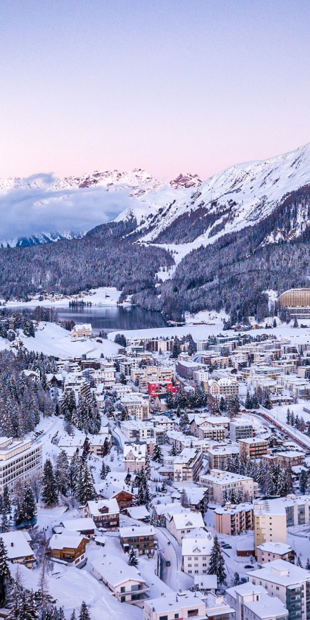 Verschneites Panorama von Davos mit dem lebendigen Alpenort, umgeben von bewaldeten Hängen, weiten Skipisten und hohen Gipfeln.