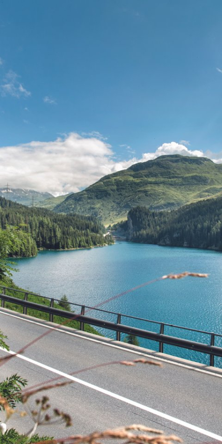 Blick auf den tiefblauen Marmorera-See am Julierpass, umgeben von bewaldeten Berghängen, Felswänden und einer kurvenreichen Passstrasse.