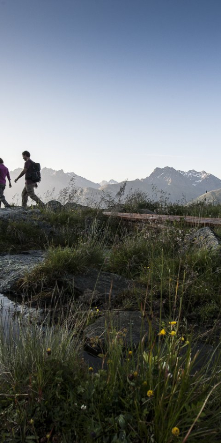 Zwei Personen wandern über einen Bergweg entlang eines kleinen Bergsees vor der eindrucksvollen Alpenlandschaft im Abendlicht.