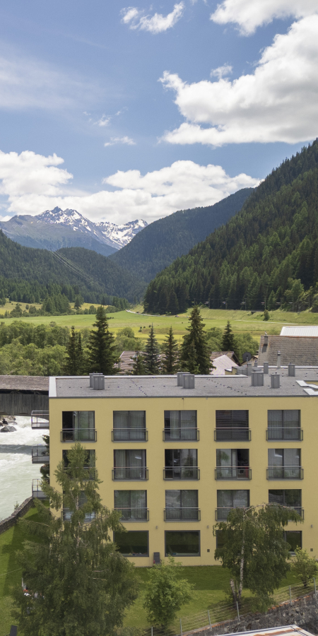 Clinica Holistica Engiadina: Modern yellow building by a river in an Alpine valley with larch forests and a snow-capped mountain.