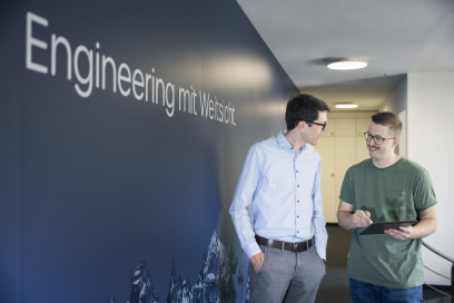 Zwei Mitarbeiter unterhalten sich im Büroflur und besprechen Inhalte auf einem Tablet vor einer Wand mit dem Schriftzug „Engineering mit Weitsicht“.