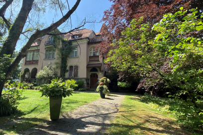 Herrschaftliches Herrenhaus mit Efeu-bewachsener Fassade, umgeben von altem Baumbestand und gepflegtem Garten mit Blumenkübeln, sonniger Tag.