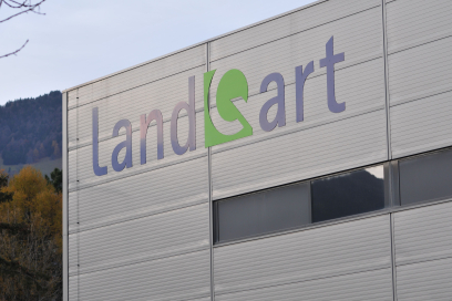 Grosses LandQart-Logo an der hellgrauen Metallfassade eines Firmengebäudes vor einer herbstlich bewaldeten Hügellandschaft.