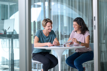 Zwei Mitarbeiterinnen bei ÖKK besprechen lächelnd Unterlagen an einem Tisch in einem hellen, modernen Büro mit Glaswänden.