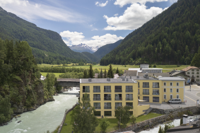 Clinica Holistica Engiadina: Modern yellow building by a river in an Alpine valley with larch forests and a snow-capped mountain.