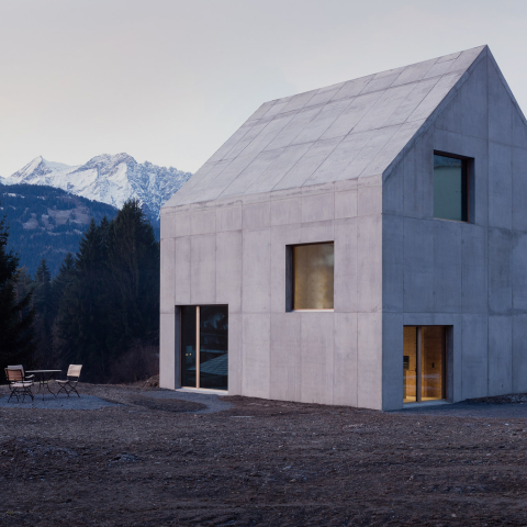 Modernes Betonhaus im Bündnerland mit markantem Giebeldach, grossen Fenstern und Terrasse vor verschneiten Bergen und dunklen Tannen.