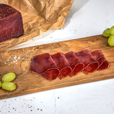 Dünn geschnittenes Bündnerfleisch auf einem rustikalen Holzplättli, angerichtet mit grünen Trauben, Salz und einem ganzen Fleischstück.