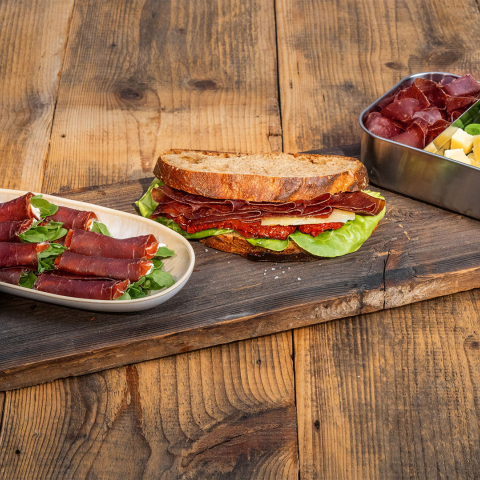 Bündnerfleisch-Plättli auf einem Holztisch mit Fleischröllchen, belegtem Brot sowie einer Box mit Käse, Nüssen und Gurken.