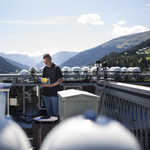Ein Forscher misst auf einem Bergdach mit technischen Sensoren die Sonneneinstrahlung, während sich hinter ihm die Bündner Alpen erstrecken.
