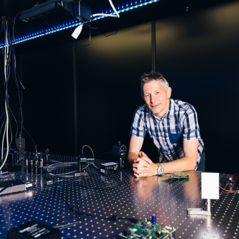 Mitarbeiter steht an einem optischen Labortisch mit Photonik-Komponenten und Präzisionsmessgeräten in einer modernen Forschungsumgebung.
