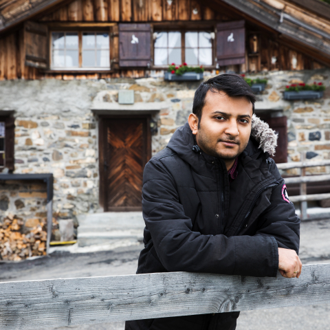 Person lehnt an einem Holzzaun vor einem traditionellen Berghaus mit Natursteinfassade und blickt direkt in die Kamera.
