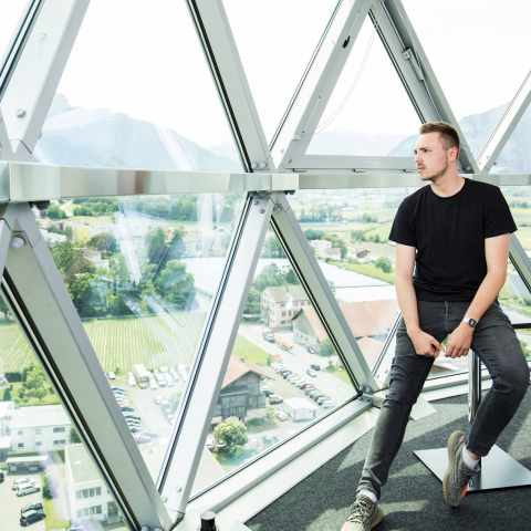 Mitarbeiter sitzt vor der grossen Glasfassade eines Aussichtsturms und blickt über das Tal mit Bergen, Feldern und Ortschaften im Hintergrund.