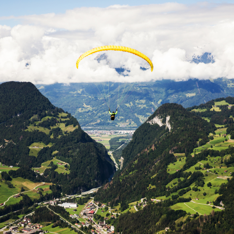 Gleitschirmflieger schwebt über der Berglandschaft Graubündens mit grünen Tälern, bewaldeten Hängen und Wolken im Hintergrund.