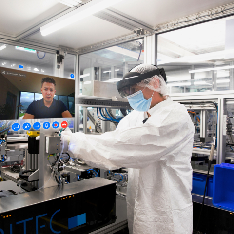 Mitarbeiter mit Schutzkleidung und Datenbrille arbeitet an einer Produktionsanlage, während ein Kollege per Videoanruf zugeschaltet ist.