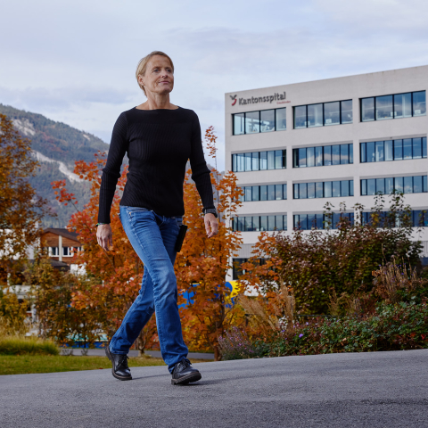 Eine Mitarbeitende des Kantonsspitals Graubünden geht durch eine herbstliche Gartenanlage vor dem modernen Spitalgebäude und der Berglandschaft.