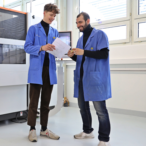 Zwei Mitarbeiter bei Polycontact besprechen technische Unterlagen in einer hellen Produktionshalle neben einer grossen industriellen Fertigungsmaschine.