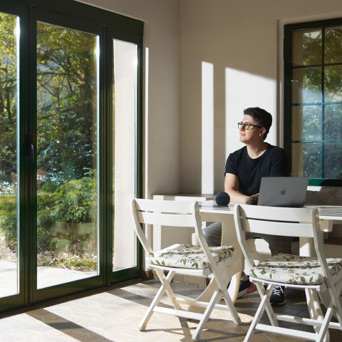 Mann mit Brille arbeitet am Laptop an hellem Schreibtisch in modernem Raum mit grossen Glasfenstern zur grünen Gartenlandschaft.