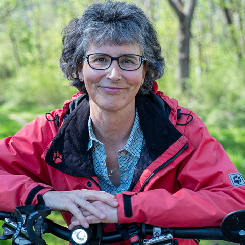 Eine Frau in roter Jacke steht für Wandel statt Wechsel mit ihrem Fahrrad auf einem grünen Waldweg und blickt lächelnd in die Kamera.