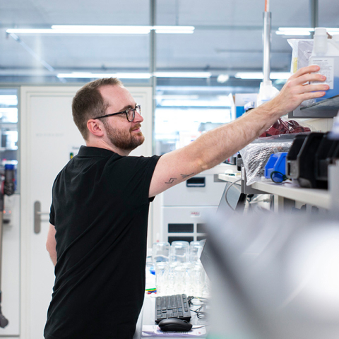 Ein Mitarbeiter bei Hamilton entnimmt in einem hellen Labor eine Probe aus einem Regal über einem Arbeitsplatz mit technischen Geräten.