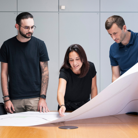 Drei Mitarbeiter bei Brüniger prüfen gemeinsam grossformatige technische Pläne auf einem Konferenztisch in einem modernen Besprechungsraum.