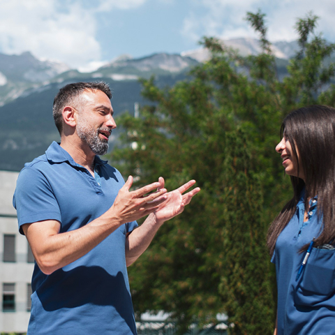 Zwei Mitarbeitende des Kantonsspitals Graubünden unterhalten sich im Freien vor dem Spitalgebäude und einer sommerlichen Berglandschaft.