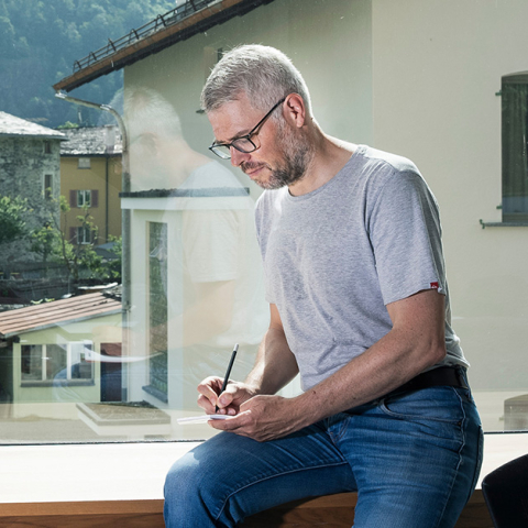 Mitarbeiter bei Repower sitzt am Fenster mit Blick auf alpines Dorf und schreibt Notizen, modernes Büro mit Berglandschaft im Hintergrund.