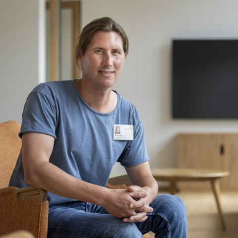 Mitarbeiter bei Psychiatrische Dienste Graubünden sitzt in modernem Wartezimmer mit braunem Sessel, grünem Sofa und Bildschirm im Hintergrund.