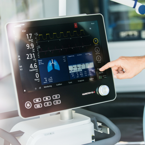 Eine Hand bedient den Touchscreen eines medizinischen Monitors mit Vitalwerten und einer Lungenanzeige an einem Beatmungsgerät.