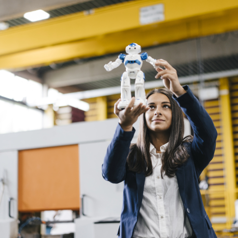 Frau hält einen kleinen humanoiden Roboter in den Händen und betrachtet ihn aufmerksam in einer modernen Produktionshalle.