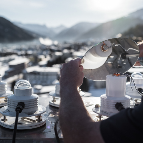 Ein Mitarbeiter justiert mehrere Messinstrumente auf einem Berg mit Blick über ein alpines Tal und die darunterliegende Ortschaft.