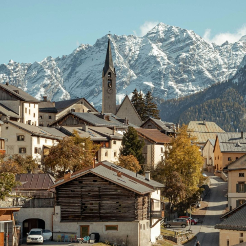 Traditionelles Engadiner Bergdorf mit Kirchturm, historischen Häusern und goldenen Lärchen vor der imposanten Kulisse schneebedeckter Gipfel.
