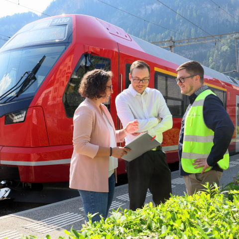 Drei Mitarbeiter der Rhätischen Bahn besprechen sich vor rotem modernem Zug in alpinem Bahnhof, Berglandschaft und Grünflächen im Hintergrund.