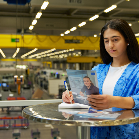 Eine Mitarbeiterin bei TRUMPF macht sich an einem Stehtisch Notizen zu einer Broschüre mit Blick auf die moderne Produktionshalle.