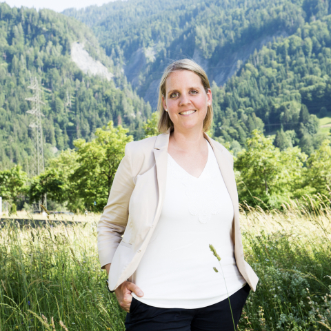 Eine Mitarbeiterin von TRUMPF steht lächelnd auf einer Wiese vor einem bewaldeten Bündner Berghang.