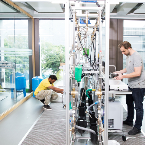 Modernes Laboratorium mit großen Fenstern: Zwei Techniker arbeiten an wissenschaftlichen Geräten und Versuchsaufbauten, helle Raumgestaltung mit Stadtblick.