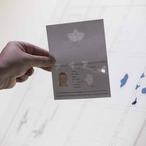 Eine Hand hält für ein Fenster in die Zukunft ein transparentes Ausweisdokument vor einen beleuchteten Bildschirm mit technischen Entwürfen.