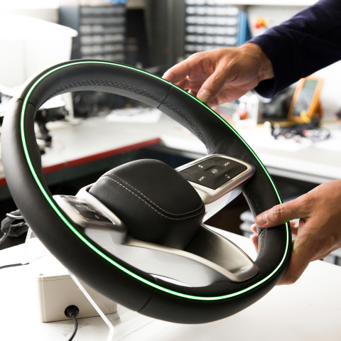Modernes Lenkrad mit grünem LED-Ring für Fahren nach Farben wird mit beiden Händen gehalten, Touch-Steuerung und Werkstatt im Hintergrund.