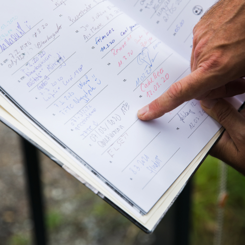 Eine Person bei LandQart zeigt auf handschriftliche Einträge und Berechnungen in einem aufgeschlagenen Logbuch im Freien.
