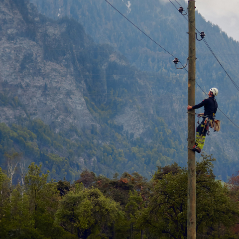 Ein Mitarbeiter von Repower klettert gesichert an einem Strommast und arbeitet an einer Leitung vor einer bewaldeten Berglandschaft.