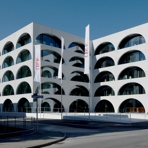 Modernes ÖKK-Gebäude mit weisser Fassade, markanten Rundbogenfenstern und Fahnen vor einer sommerlichen Bündner Berglandschaft.