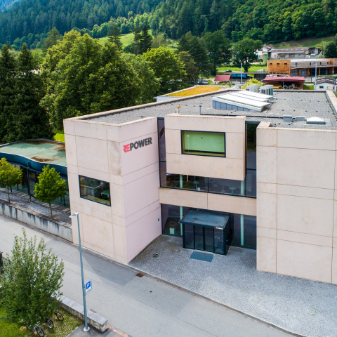 Repower-Gebäude mit grauem Betonbau und großem Logo in alpinem Tal, umgeben von dichten Fichtenwäldern, Fluss und Berglandschaft im Hintergrund.