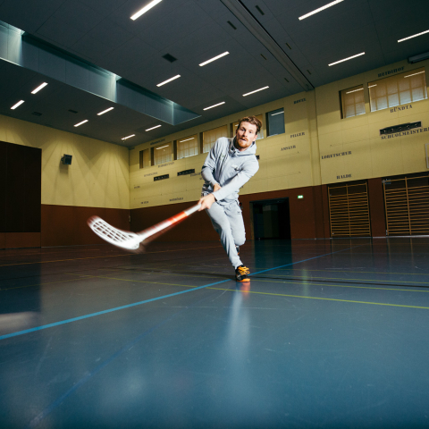 Ein Hockeyspieler schiesst mit einem Unihockeystock einen weissen Ball durch eine grosse, dunkel beleuchtete Sporthalle.
