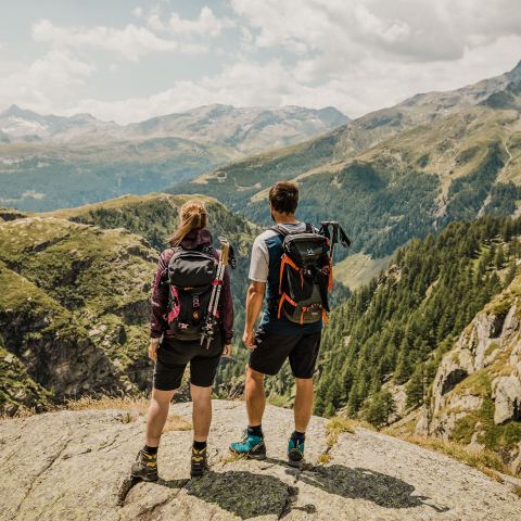 Zwei Bergsteiger stehen mit Rucksäcken auf einem Gipfel und blicken über ein weites alpines Bergpanorama mit Felsen und Wäldern.