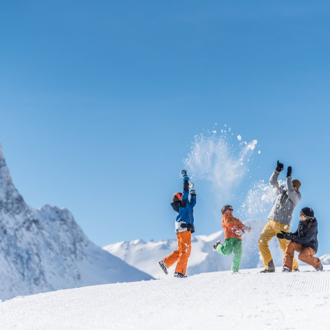 Eine Familie wirft auf einer verschneiten Piste jubelnd Schnee in die Luft, umgeben von eindrucksvollen Gipfeln unter blauem Winterhimmel.