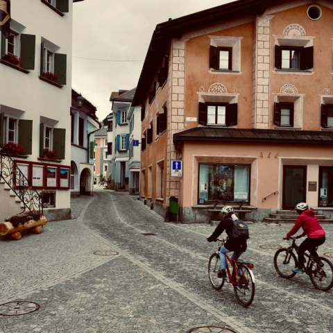Zwei Velofahrende fahren durch die gepflasterte Gasse eines Engadiner Dorfs in der Region Maloja, vorbei an Häusern mit Sgraffiti-Fassaden.