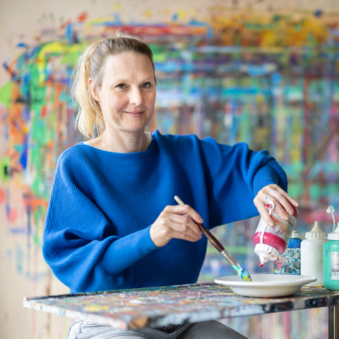 Frau bei Psychiatrische Dienste Graubünden malt mit Pinsel und Farben an Kunstwerk, bunte abstrakte Gemälde im Hintergrund, therapeutische Kunstaktivität.